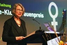 Foto: Eine Frau mit kurzen, grauen Haaren steht an einem Rednerpult und hält ein Blatt Papier. Im Hintergrund ist ein Bildschirm mit Text und Symbol.