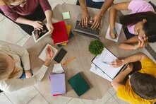 Foto: Fünf Personen sitzen an einem Tisch mit Laptop, Tablet, Notizen und Büchern, arbeiten gemeinsam an einem Projekt.