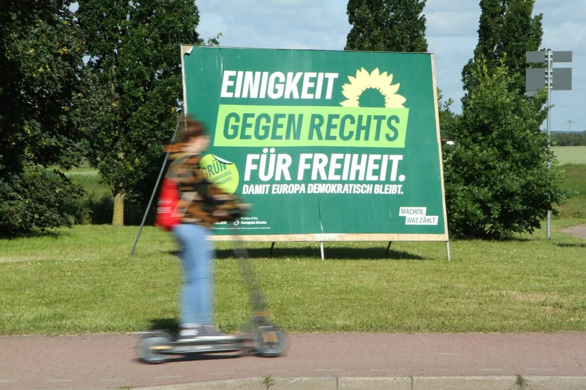 Wahlplakat der Grünen mit dem Schriftzug Gegen Rechts im Mittelpunkt. Im Vordergrund fährt eine Person auf einem Roller vorbei.